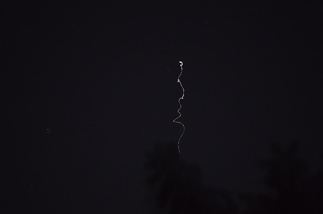 A dark night sky with a thin, elongated silhouette resembling a human face formed by light trails.